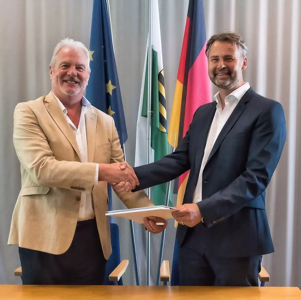 Two men shake hands over the signed licence agreement, in front of flags at Fraunhofer IKTS, Dresden, July 2019